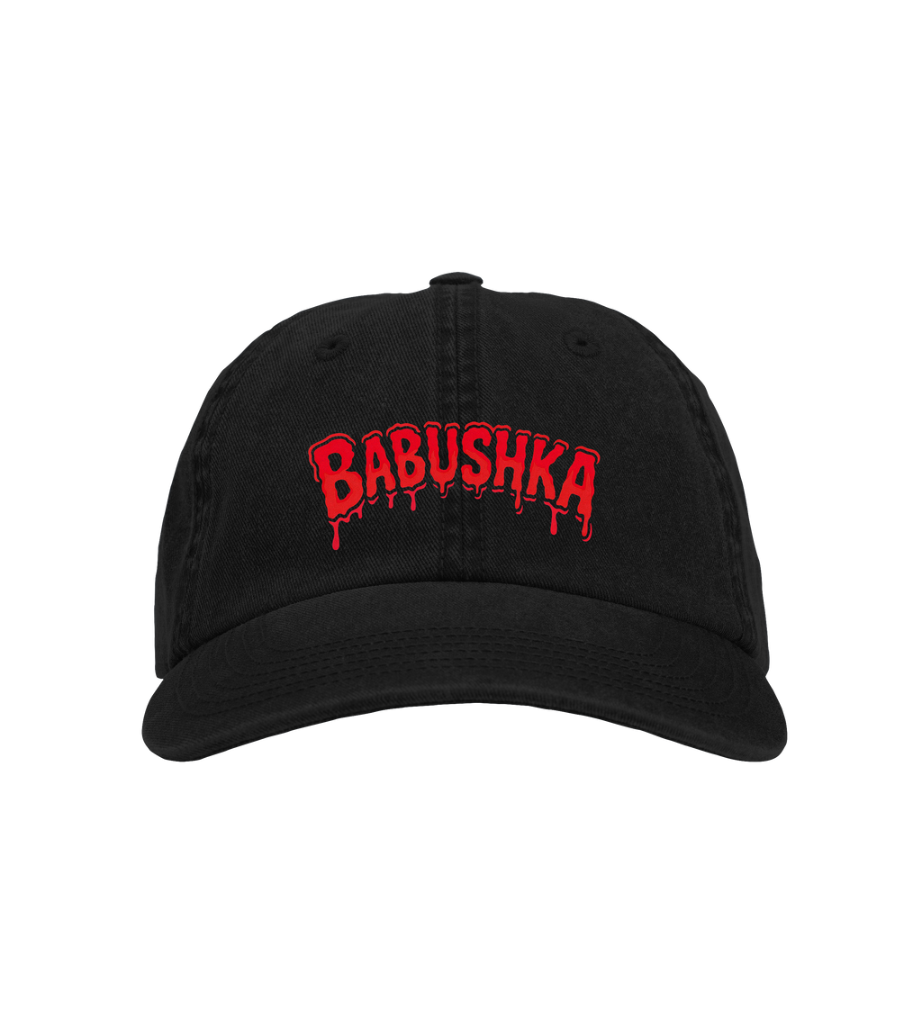 Black six-panel organic cotton baseball cap with red embroidered “BABUSHKA” logo, hard brim, velcro strap, and ridged texture.